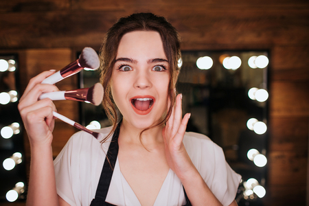 Emotional amazed young woman look on camera in beauty room she hold set of eye brushes for make up mirror with light bulbs behind