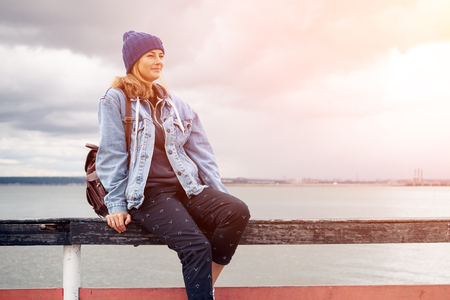 Outdoor atmospheric lifestyle photo of young beautiful darkhaired woman in knitting hat in a denim jacket and black trousers relaxing and sitting against the background sea in sunny autumn day