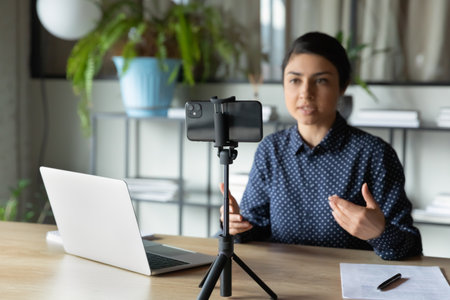 Serious indian presenter girl speaker talking at webcam on smartphone online teacher tutor coach giving webinar from home woman using mobile phone on tripod for remote online conference call