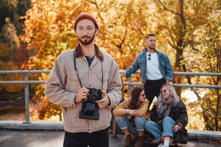 Bearded and warm dressed guy with glasses posing in foreground in background of his friends and autumn landscape in park Stock Photo