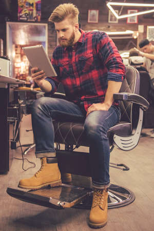 Handsome bearded man is using a digital tablet while sitting in chair at the barbershop Stock Photo