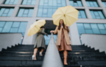 Two stylish women ascending steps outside a modern building holding vibrant yellow umbrellas on a possibly rainy day