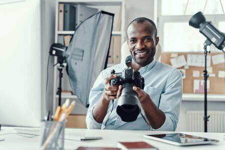 Handsome young african man holding digital camera and smiling while working in the modern office Stock Photo