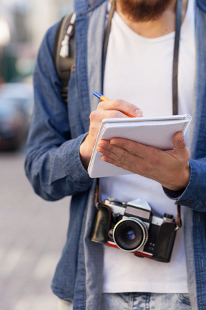 Close up of hands of explorer making journey in city he is standing and writing down some notes the man is carrying backpack and camera