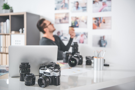 Focus on camera and notebook computer locating on table side view serene bearded man looking at photos on wall while tasting mug of coffee profession concept Stock Photo