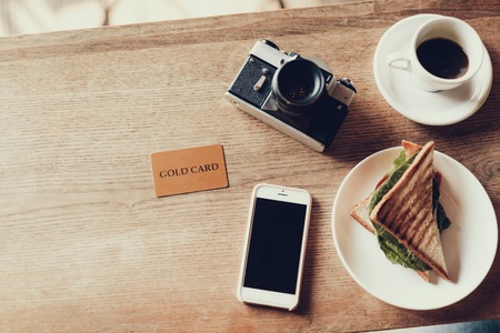 Flat lay photo of gold plastic card photo camera cellular and beverage with sandwich lying on wooden desk