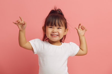 Joyful three years old asian ethnicity little girl raises her hands up expresses a cheerful mood smiles happily standing on a pink isolated Foto de archivo