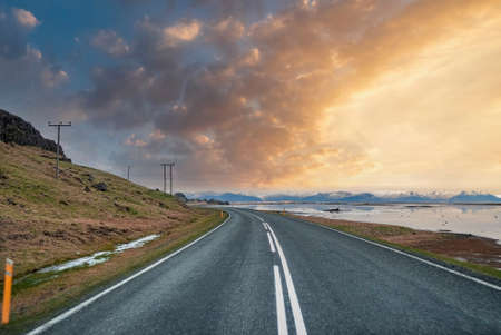 Diminishing highway amidst sea and mountain against cloudy sky during sunset