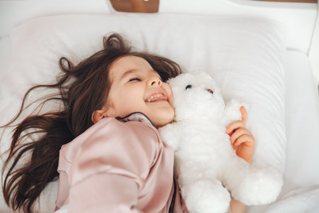 A little dark haired girl is sleeping in bed hugging a teddy bear under a white blanket the child is in bed in the morning