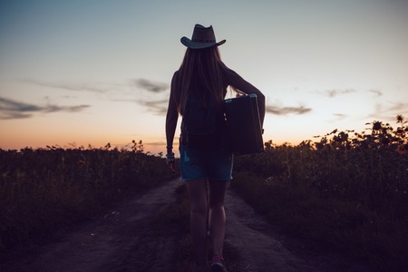 Girl in a cowboy hat standing with a suitcase on the road in the sunflower field waiting for help sunset