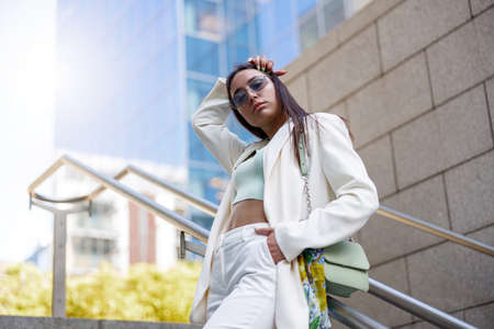 Fashionable woman in white suit standing on stairs on background of modern buildings
