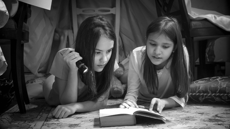 Black and white portrait of two teenage girls lying on floor and reading book with flashlight Stock Photo