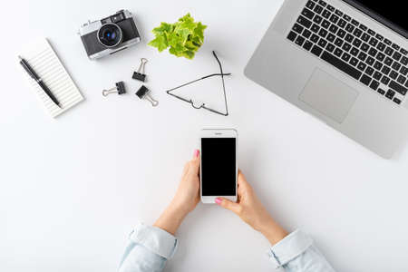 Overhead shot of young woman using smart phone over white desktop with laptop and business accessories flat lay