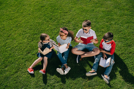 Multiethnic children in sunglasses reading books on green meadow Stock Photo