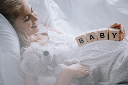 Side view of smiling pregnant woman in white nightie with teddy bear and wooden blocks with baby lettering on belly resting on sofa