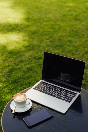 Laptop and smartphone with blank screens and cup of cappuccino on table in garden
