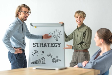 Laughing businessmen showing strategy inscription and business signs on flipchat to businesswoman