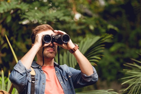 Adult traveler looking through binoculars in tropical green forest