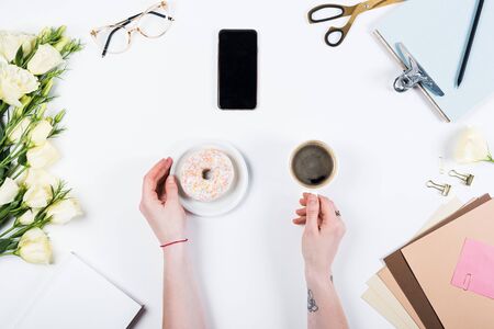 Cropped view of woman with doughnut and cup of coffee at workplace