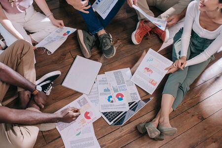 Cropped view of young businesspeople sitting on floor near documents with infographics