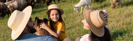 Girl in straw hat smiling near cattle dog and parents on green pasture banner Foto de archivo