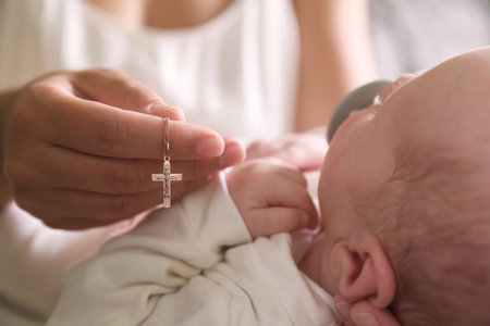 Mother holding christian cross near newborn baby indoors focus on hand