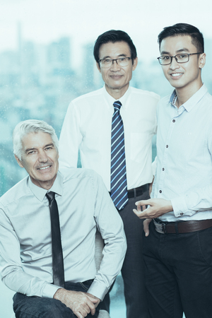 Three smiling multiethnic businessmen looking at camera senior caucasian man sitting on chair his asian coworkers standing near him team concept Stock Photo