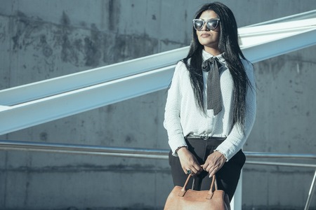 Determined young businesswoman in sunglasses looking into distance and holding bag outdoors