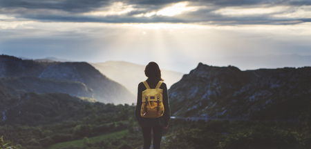 Hipster young girl with bright backpack enjoying sunset on peak of foggy mountain