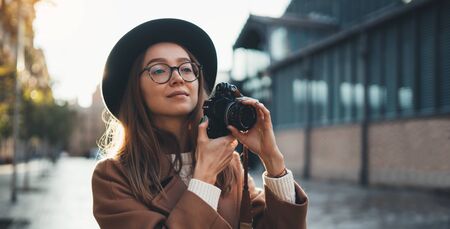 Hobby photographer concept outdoor lifestyle portrait of pretty woman in sun city in europe with camera travel photo of photographer in glasses and hat take photo copy space mockup