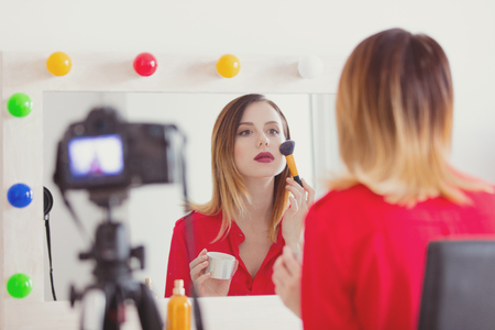 Young caucasian blogger woman applying cosmetics at camera for video sharing website home location near a mirror