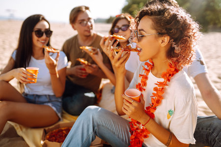 Grupo de jóvenes amigos haciendo picnic, comiendo pizza, brindando con cerveza en la playa.