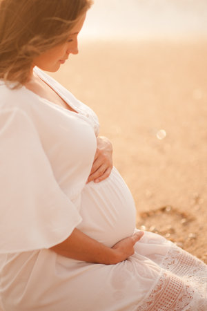 Pregnant woman wearing white dress sittin over sea shore outdoors motherhood maternity healthy lifestyle Фото со стока