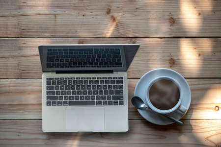 White ceramic coffee cup and computer note book on wooden table with morning nature light with copy space