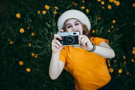 Young beautiful girl in an orange t shirt holding retro camera in her hands lying on the lawn where dandelions grow flowering dandelions