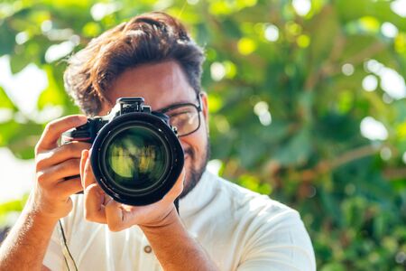 Handsome and confident indian man photographer with a large professional camera taking pictures photo shooting on the beach photo session on summer holiday on the background of green tropical trees