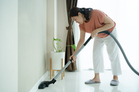 Joven ama de llaves asiática que usa una máquina de vacío para limpiar un piso sucio en la sala de estar cerca con espacio de copia. Ama de casa aspirando un piso y un sofá en la sala de estar. Foto de archivo