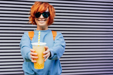 Glass of fruity sugar flavored tapioca pearl bubble tea with straw in hand of hipster fashion woman in heart sunglasses and blue clothes on gray striped wall background selective focus copy space