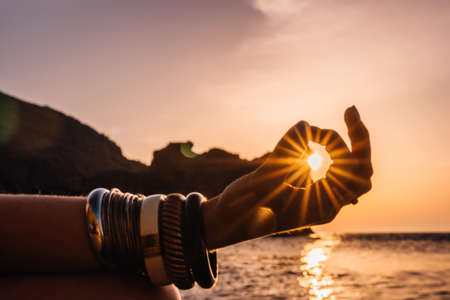 Young woman with long hair in white swimsuit and boho style braclets practicing outdoors on yoga mat by the sea on a sunset womens yoga fitness routine healthy lifestyle harmony and meditation