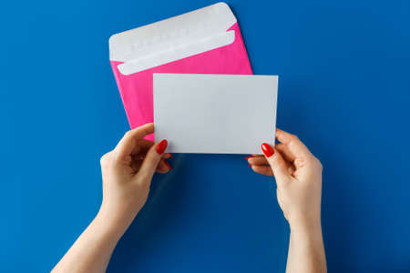 Rosa Umschlag mit einer leeren Karte in den Händen auf blauem Hintergrund. Hände halten einen rosa Umschlag mit einem leeren Buchstaben auf blauem Hintergrund. Standard-Bild