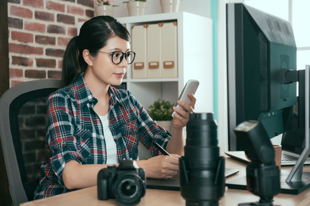 Successful confidence female photographer sitting in office desk editing photo and using mobile smartphone checking picture seriously