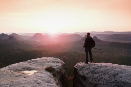 Tall tourist guide on rock with pole in hand hiker with sporty backpack stand on cliff above misty valley sunny spring daybreak in rocky mountains