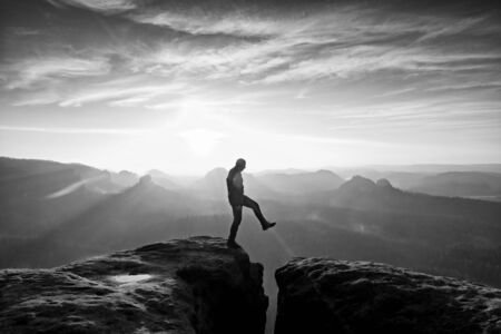 Man in black jumping between rocky empires dreamy daybreak in sandstone mountains heavy orange mist in deep valley