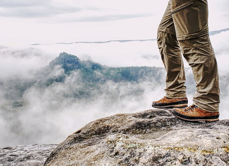 Long tired male legs in hiking trousers stay for moment on peak of rock above valley Stock Photo