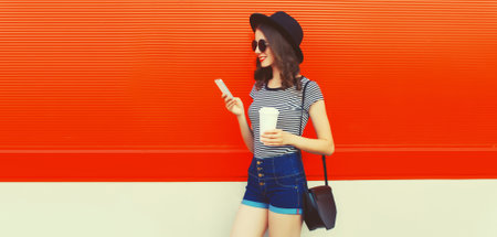 Portrait of young woman with smartphone and cup of coffee wearing black round hat on orange background