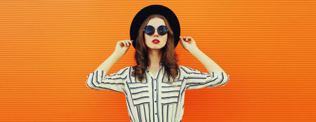 Portrait of beautiful young woman posing wearing white striped shirt black round hat on orange background