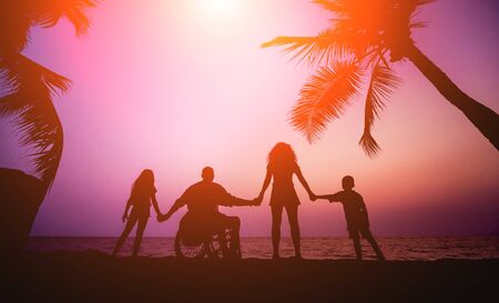 Hombre discapacitado en silla de ruedas con su familia en la playa. siluetas al atardecer Foto de archivo