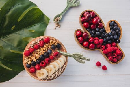 Acai smoothie granola seeds fresh fruits in a wooden bowl with cactus spoon plate filled with berries on white wooden background with with monstera leaf Stock Photo