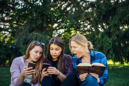 Three cute girls relax and socialize on the lawn in the summer park young women on the green grass among the trees looking at smartphone reading a book students in between classes outdoor