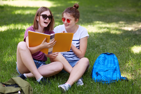 Horizontal shot of happy female studens feels relaxed while sit crossed legs on green grass enjoy spring weather have positive expressions rests after classes people rest leisure concept